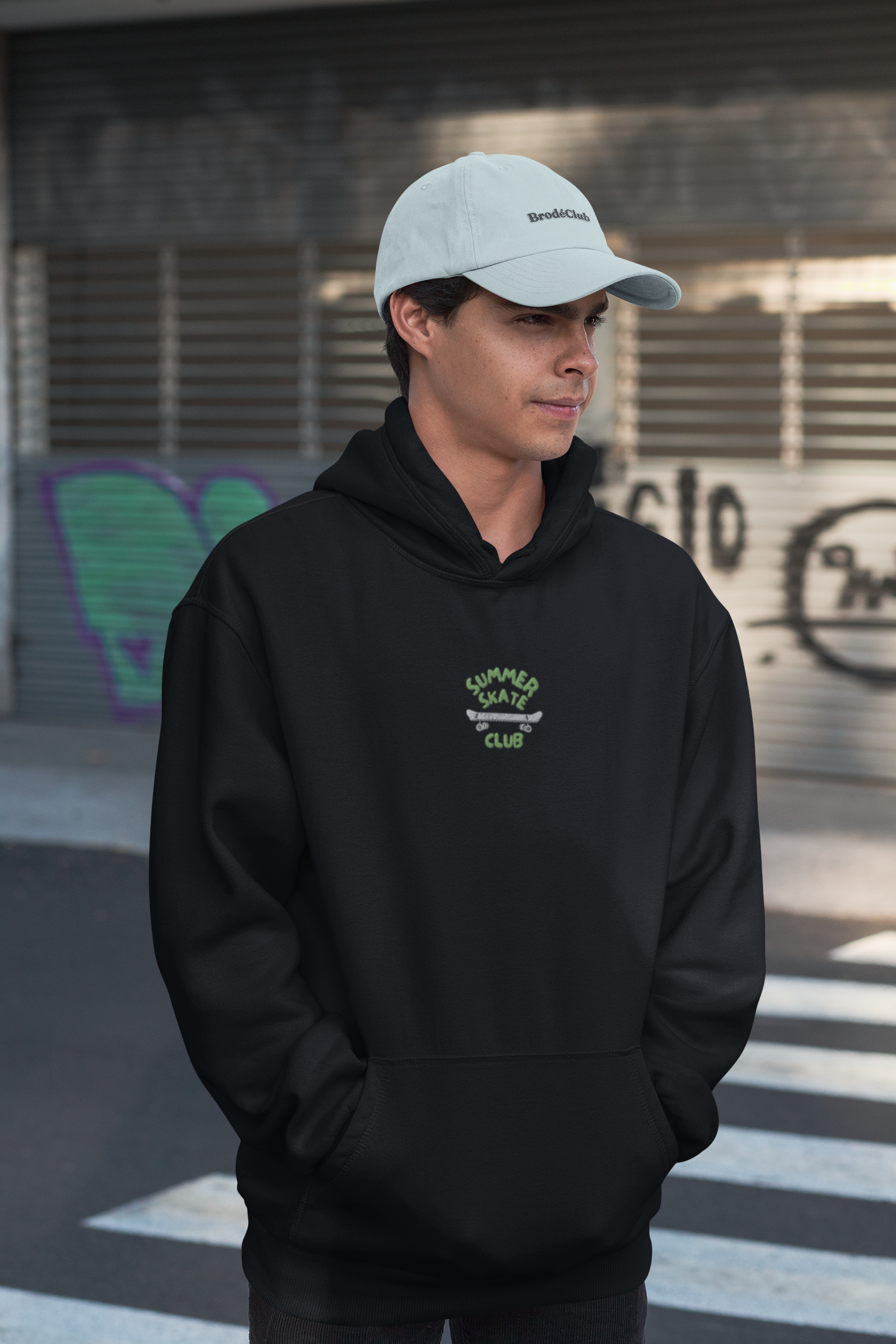 Person wearing a black hoodie with a logo and a white cap, standing in an urban setting.