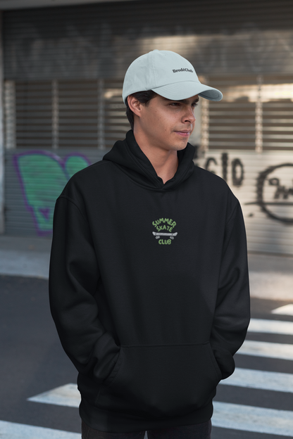 Person wearing a black hoodie with a logo and a white cap, standing in an urban setting.